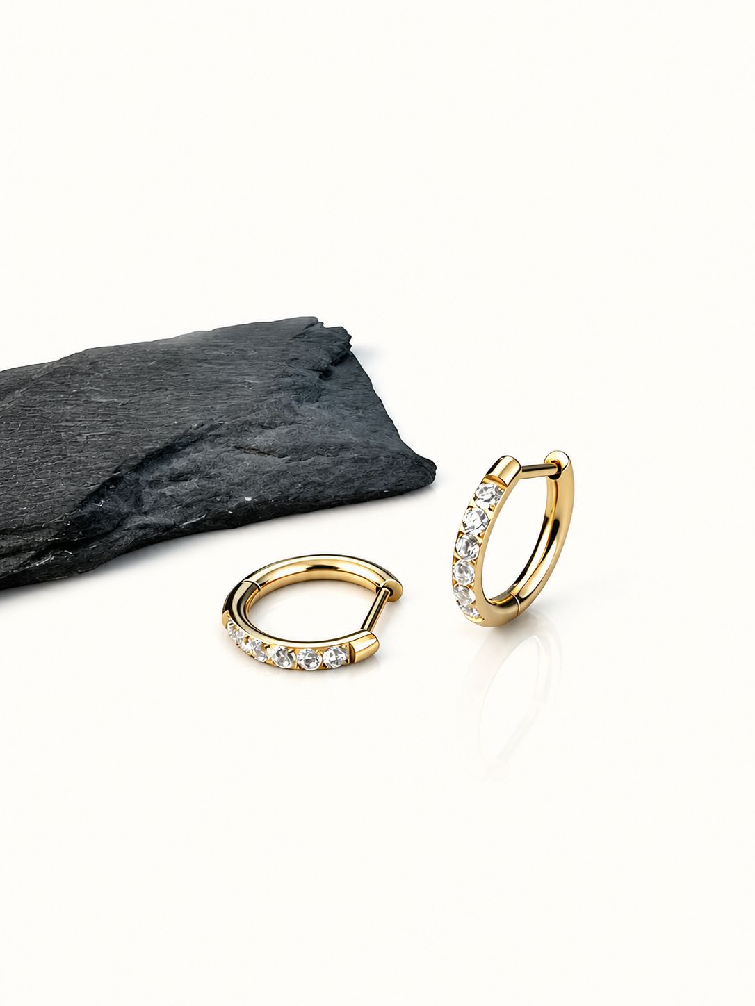 Two gold G23 titanium pavé CZ clicker hoops styled beside a black stone on a neutral background.
